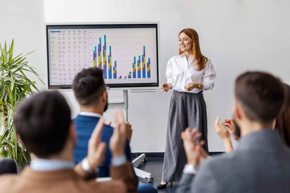Portrait of data expert standing by panel with data analysis and having speech in front of her team at corporate office.